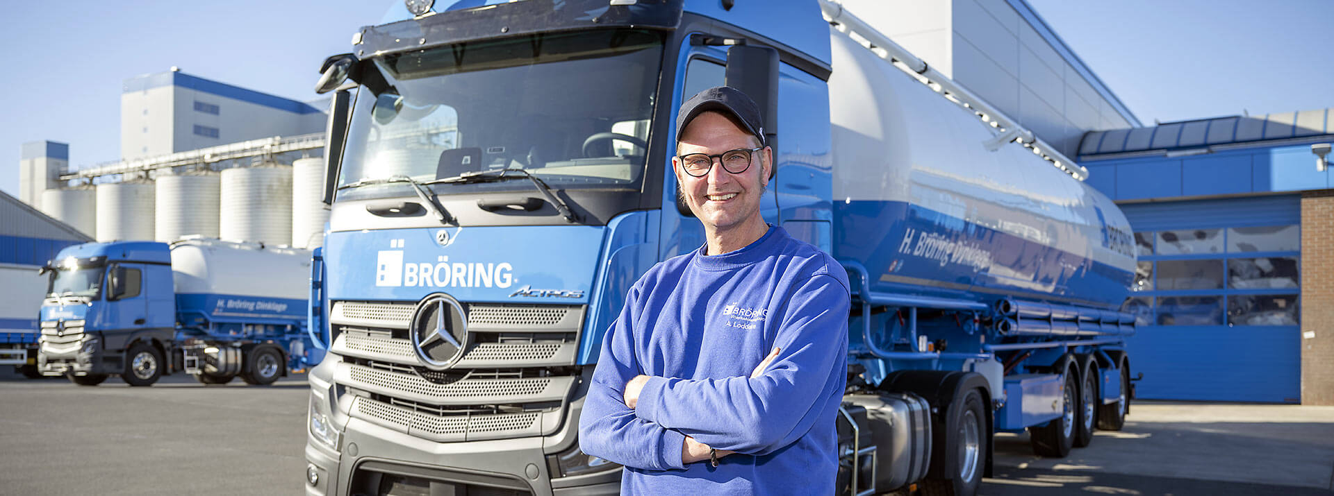 LKW-Fahrer steht vor BRÖRING Silo-LKW, lächelt freundlich in die Kamera. Im Hintergrund Futtermittelproduktionswerk und Waschanlage sowie weiter LKWs